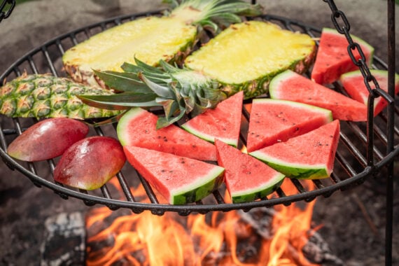 Slices of pineapple, watermelon, and mango are arranged on a round grill suspended above an open flame, with the fruits being grilled outdoors.