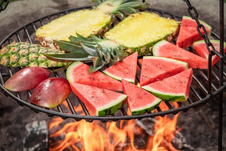 Slices of pineapple, watermelon, and mango are arranged on a round grill suspended above an open flame, with the fruits being grilled outdoors.