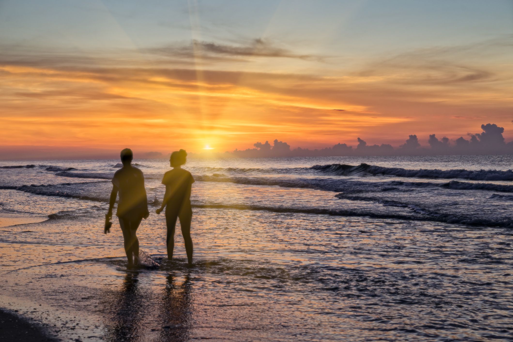Male and female couple walking in ocean along beach of Myrtle Beach, South Carolina during dramatic early sunrise, purple ocean and sky with bright orange and yellow sky