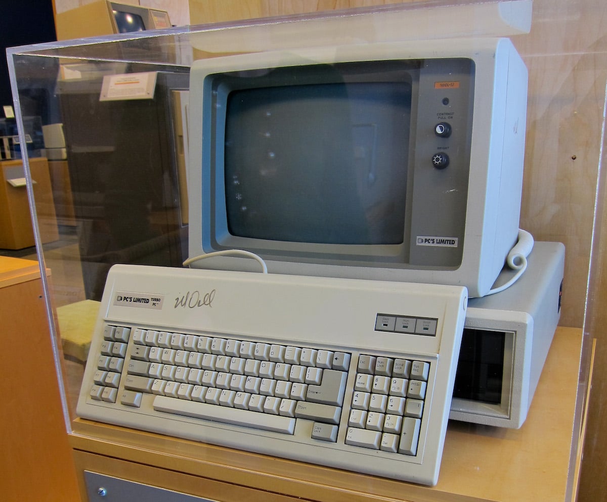 A vintage DK'S Limited personal computer with a bulky CRT monitor, keyboard, and external floppy disk drive, reminiscent of companies founded by year, all displayed inside a clear protective case on a wooden surface.