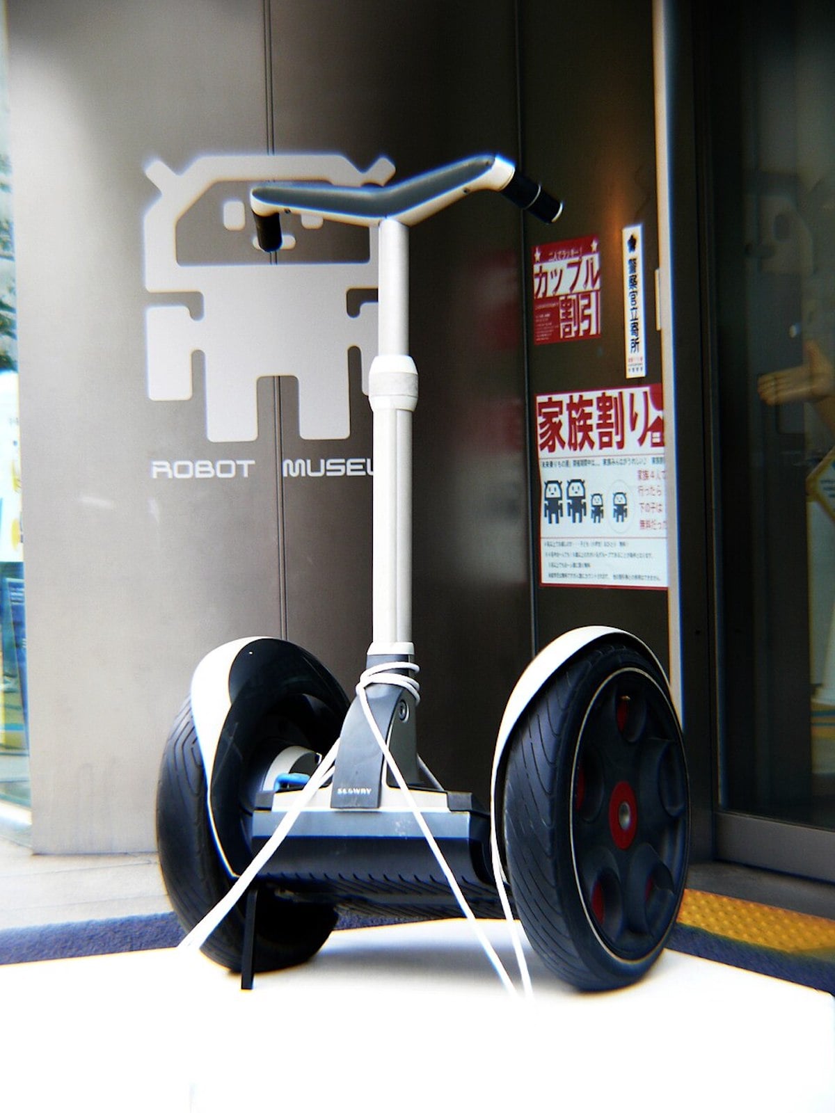 A Segway personal transporter is parked outside a building with a "Robot Museum" sign and logo on the wall, highlighting innovations by companies founded by year. Posters and glass doors are visible in the background.