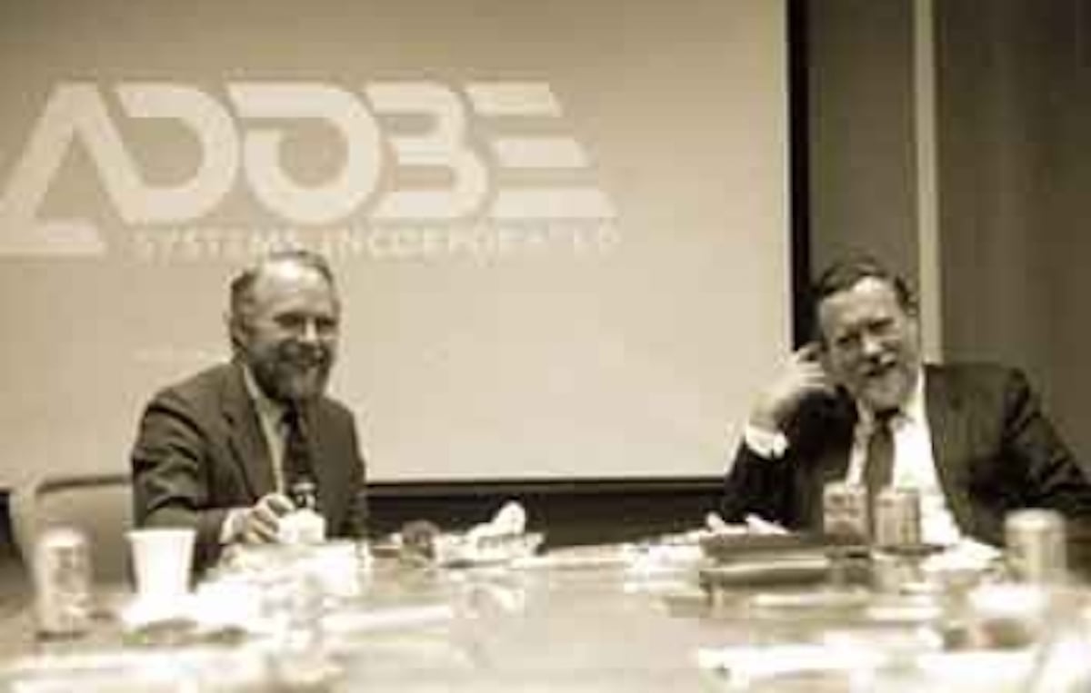 Two men in suits sit at a table with drinks and papers, smiling, in front of a screen displaying the Adobe Systems Incorporated logo—one of the notable companies founded by year. The scene appears to be a business meeting or discussion.