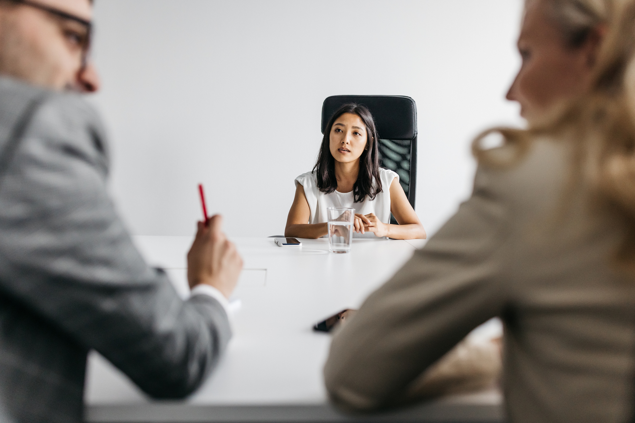 Young woman having a job interview sitting at one end of a white table in the background with a male and female interviewer blurred in the foreground, a clean white wall in the background