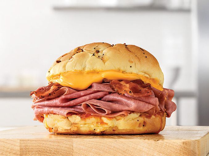 A sandwich with layers of sliced roast beef, crispy bacon, and melted yellow cheese on a toasted onion bun, placed on a wooden surface with a blurred kitchen background.