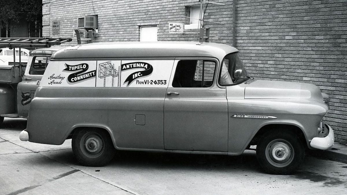 Black-and-white photo of a vintage van parked by a brick building. The van features "Tupelo Community Antenna, Inc.," one of the companies founded by year, with a phone number promoting its antenna and TV service. Comcast 1963