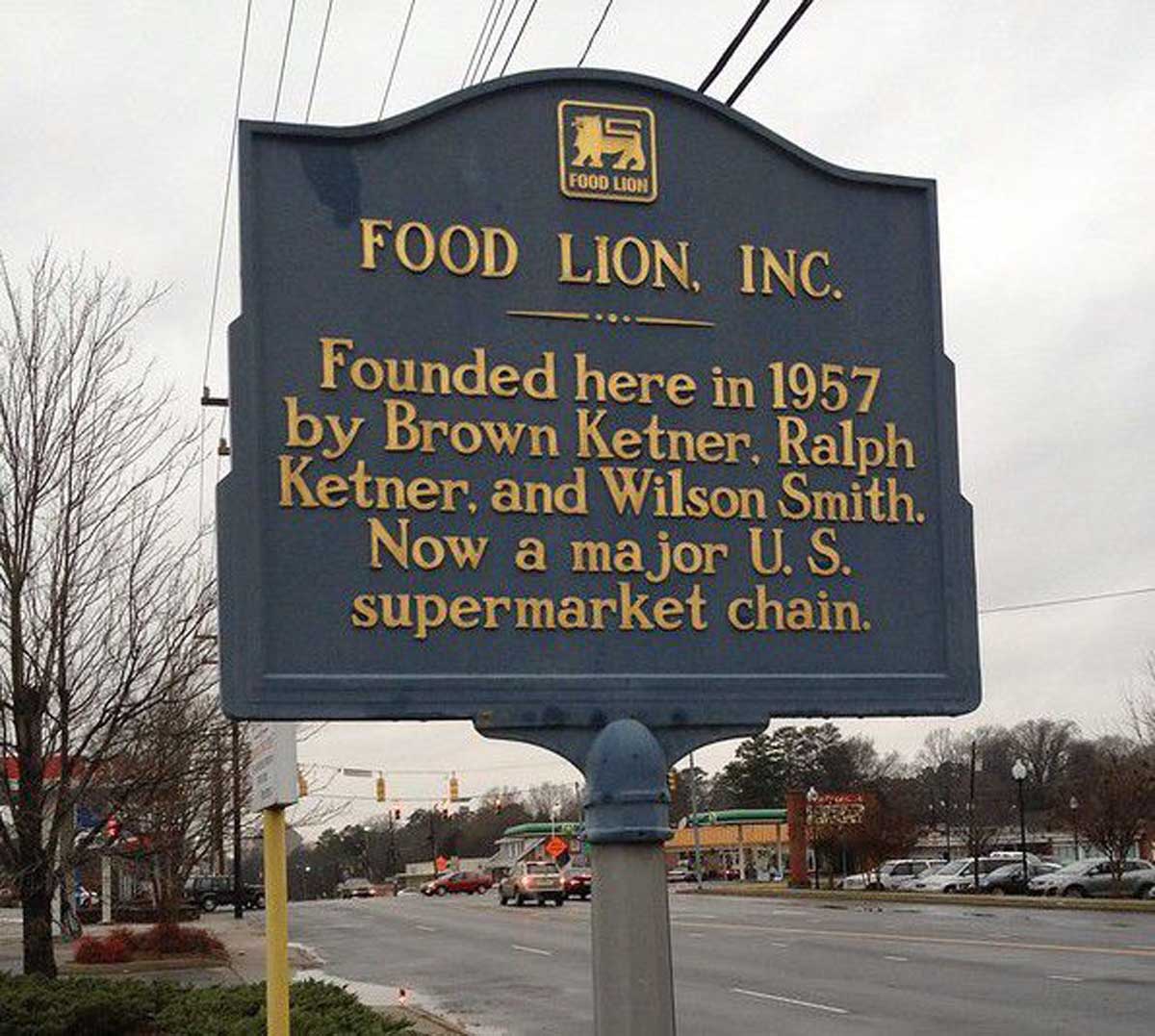 A blue historical marker sign for Food Lion, Inc., one of the companies founded by year in 1957 by Brown Ketner, Ralph Ketner, and Wilson Smith; now a major U.S. supermarket chain. The sign is on a street with cars and trees visible.