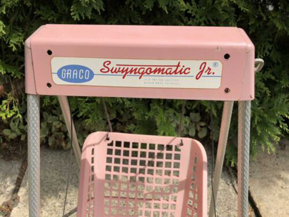 A close-up of a vintage pink Graco Swingomatic Jr. baby swing, from one of the companies founded by year, shows the label on the top bar with trees and greenery in the background.
