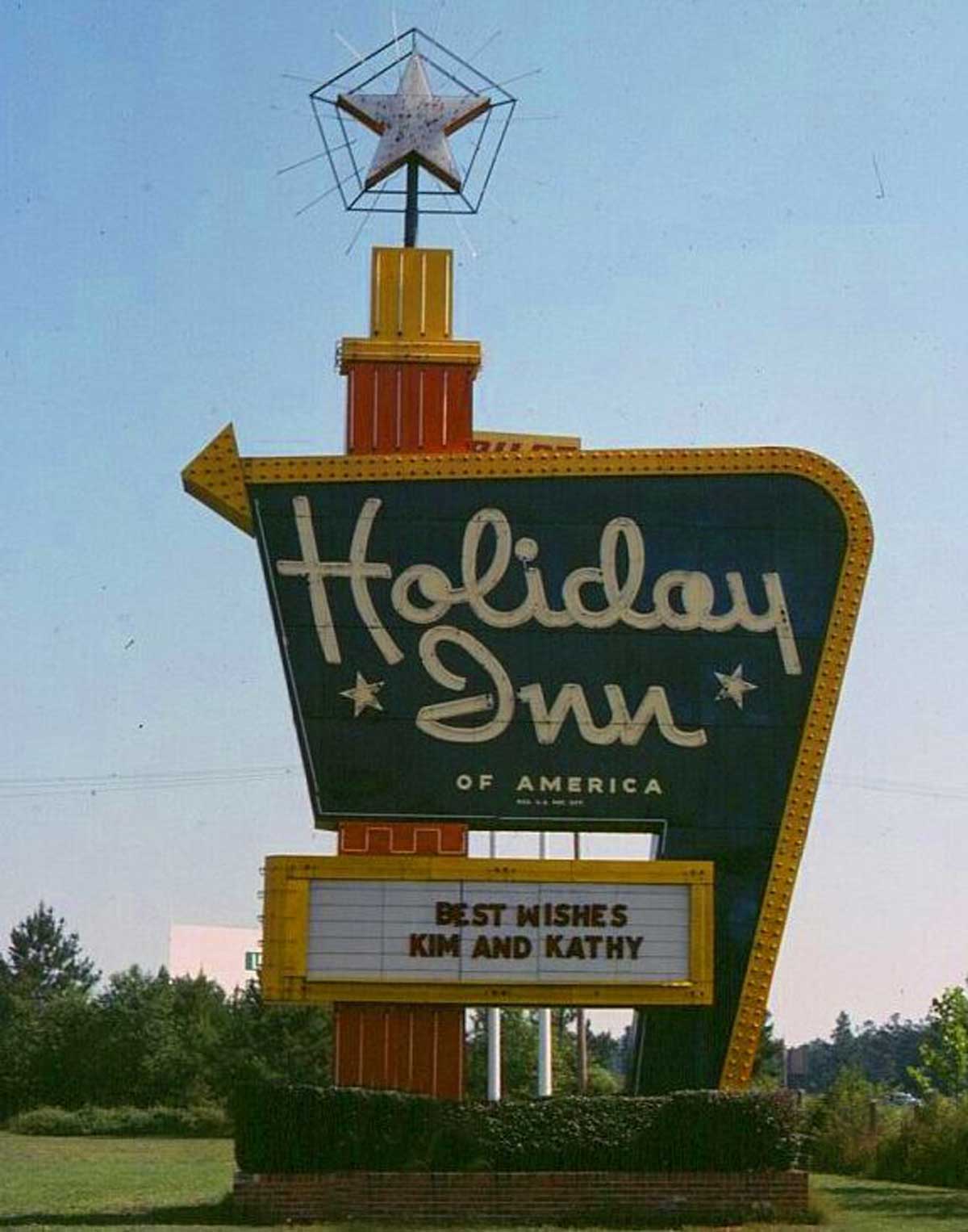 A retro Holiday Inn of America sign, representing one of the iconic companies founded by year, stands outdoors with a star on top. The marquee below reads "BEST WISHES KIM AND KATHY." Trees and grass are visible in the background under a clear sky.