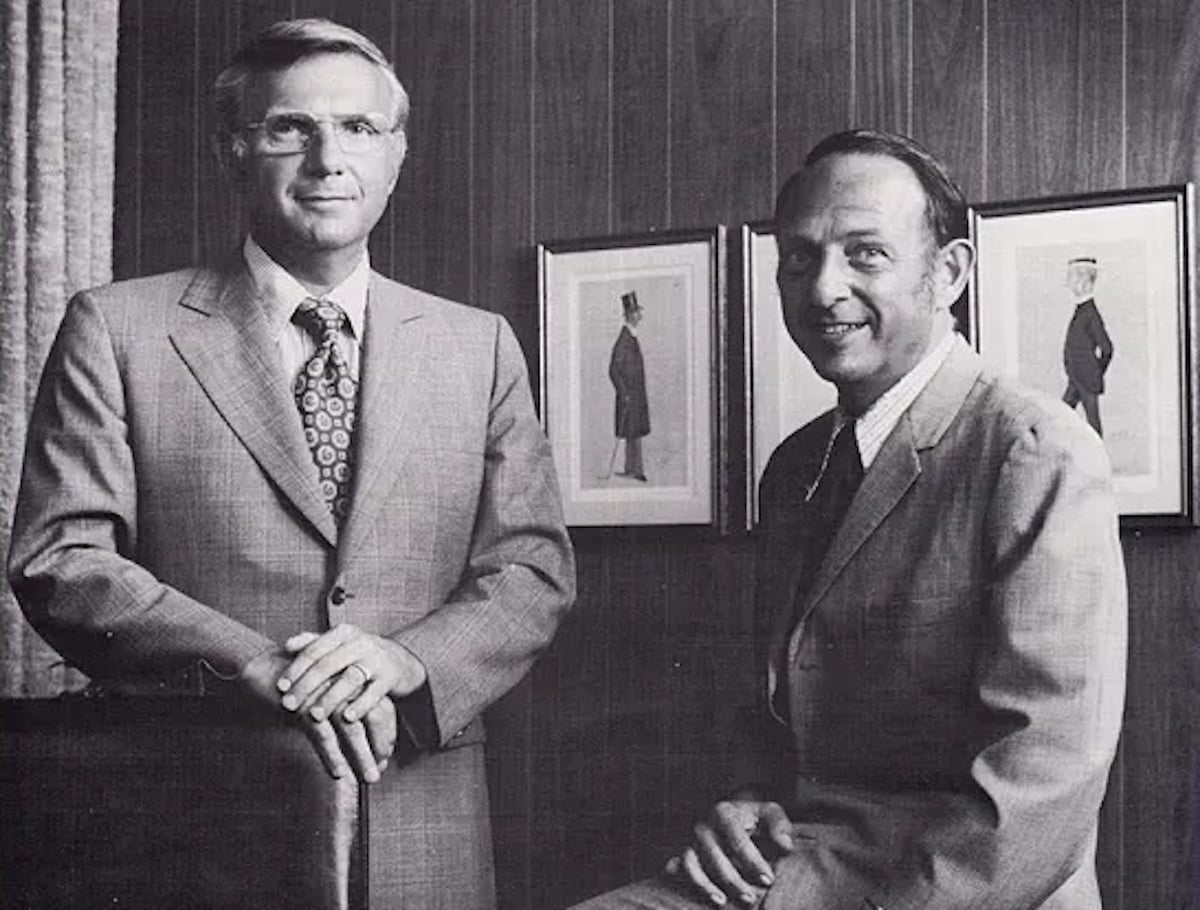 Two men in suits pose in an office with wood-paneled walls and framed artwork—an atmosphere reminiscent of classic companies founded by year. One stands with his hand on a chair, the other sits, both facing the camera and smiling slightly.