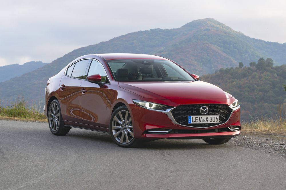 A red Mazda sedan is parked on a road with green hills and mountains in the background under a cloudy sky.