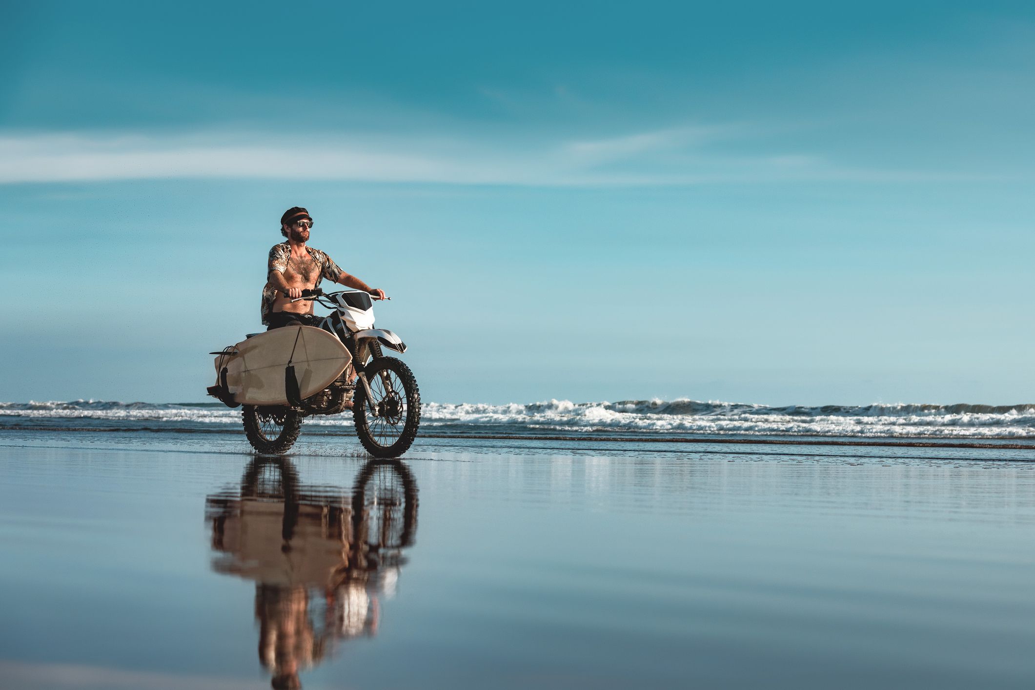 male surfer rides on motorcycle with surfboard by ocean beach