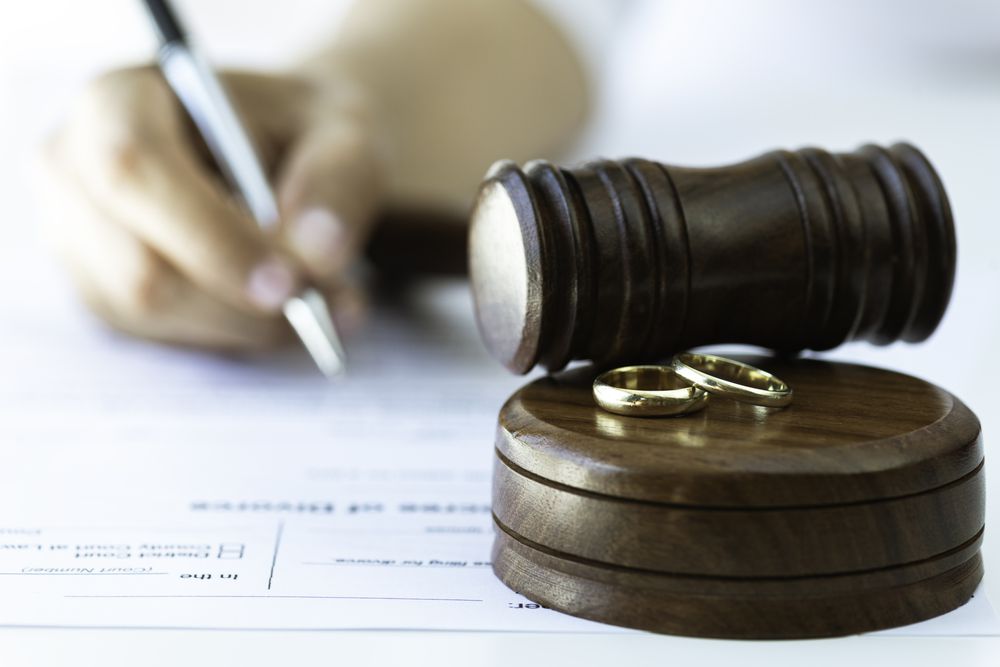 Close-up of two wedding rings and a judge’s gavel on a desk, with a person in the background signing a document, suggesting themes of divorce, legal separation, or marriage law.