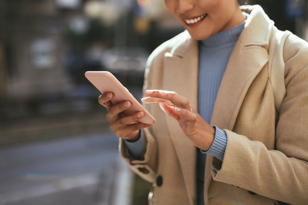 A person wearing a beige coat and blue turtleneck sweater is smiling while using a smartphone outdoors. The background is blurred, focusing attention on their hands and phone.