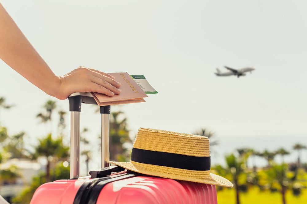 A person rests a hand with a passport and boarding pass on top of a pink suitcase with a straw hat on it, while an airplane takes off in the background amid palm trees.