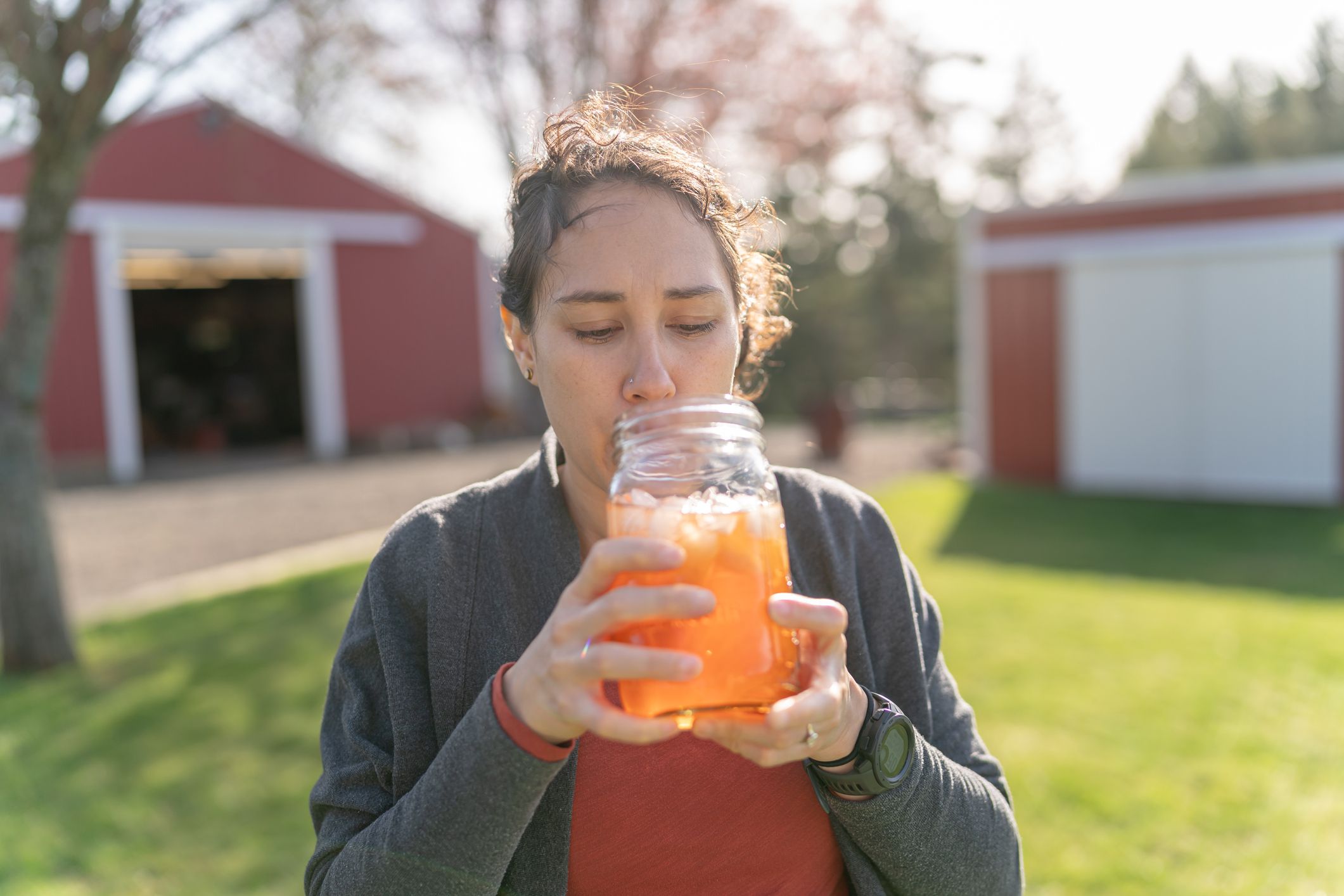 A multiracial woman stands outside in the backyard and drinks ice tea from a mason jar on a warm and sunny spring day. A red barn and trees are visible in the background.