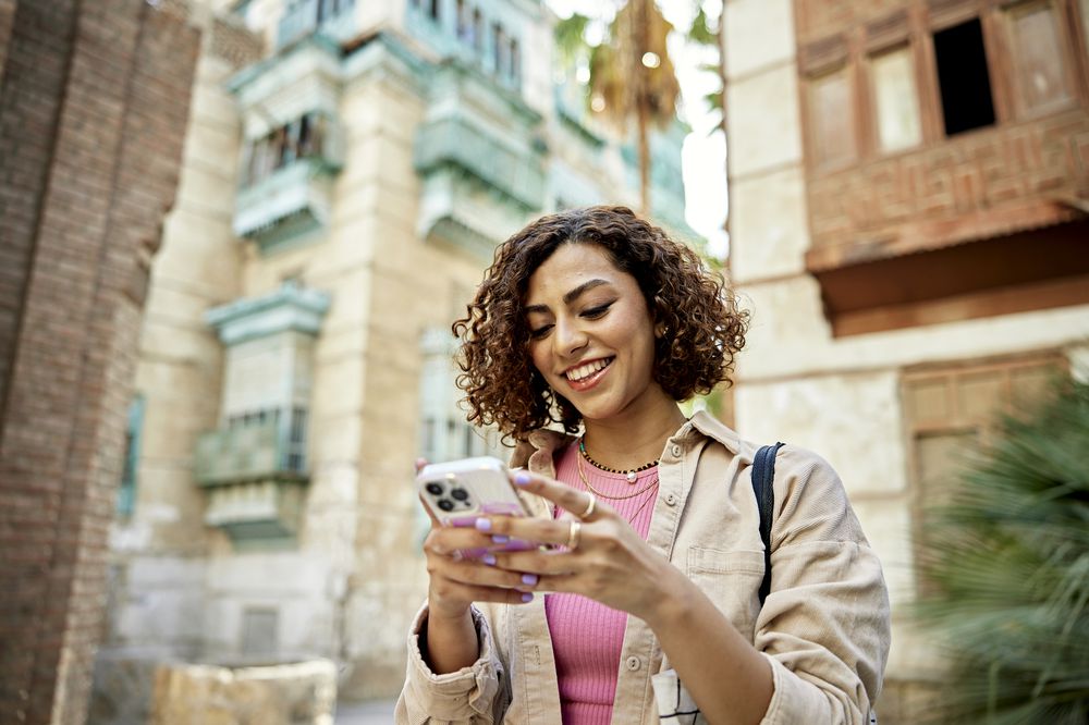 A woman with curly hair smiles while using her smartphone outdoors, standing in front of historic buildings with intricate architectural details.