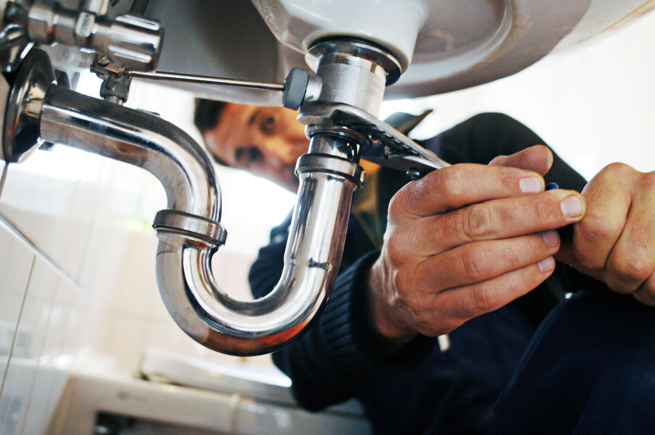 A person uses a wrench to tighten a pipe under a sink, focusing on plumbing repairs. The image shows hands and tools close-up, with the plumber's face blurred in the background.