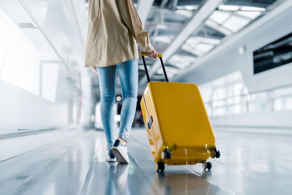 A person in jeans and a tan jacket walks through an airport terminal, pulling a bright yellow suitcase on wheels along a moving walkway. The setting is modern and brightly lit.