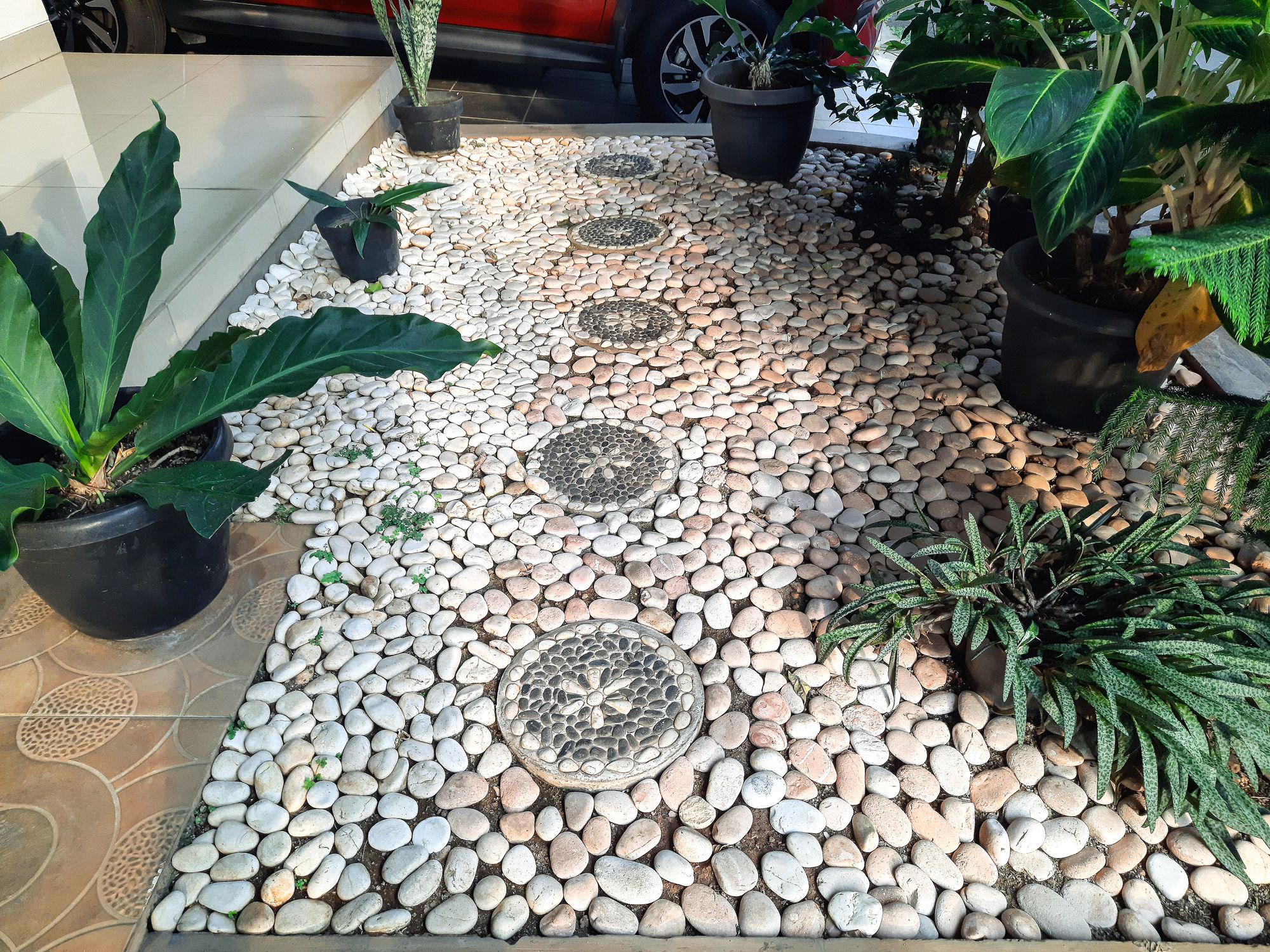 View of garden in front of a house. Garden with pebbles and green plants in pots. A red car parked in the carport beside the garden.