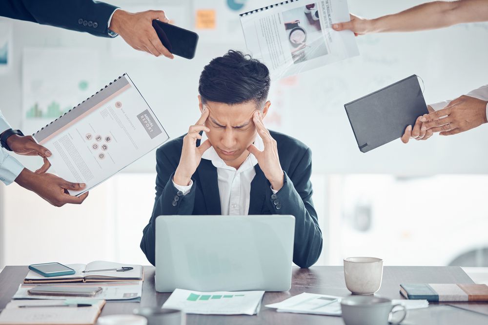 A stressed man in a suit sits at a desk with a laptop, holding his head as several hands reach in, offering documents, a phone, and a tablet, suggesting he is overwhelmed by work.