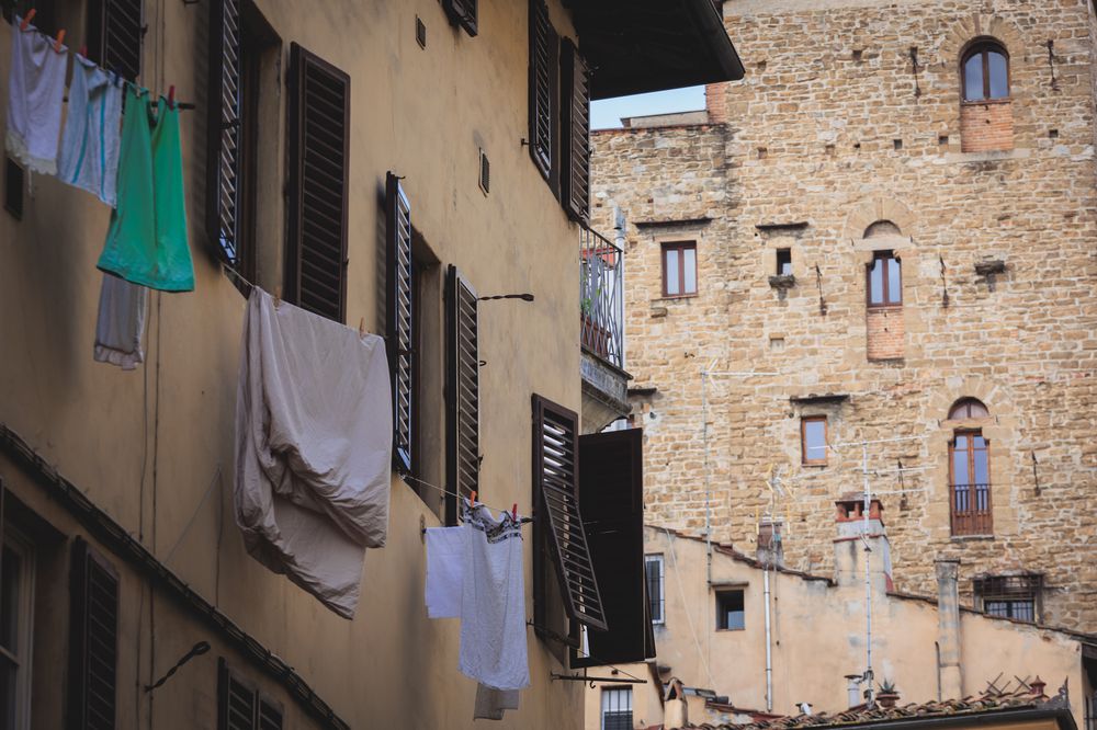 Laundry hangs on clotheslines strung between windows of an old, yellow-stucco building with wooden shutters, set against a background of a historic stone building with small windows.