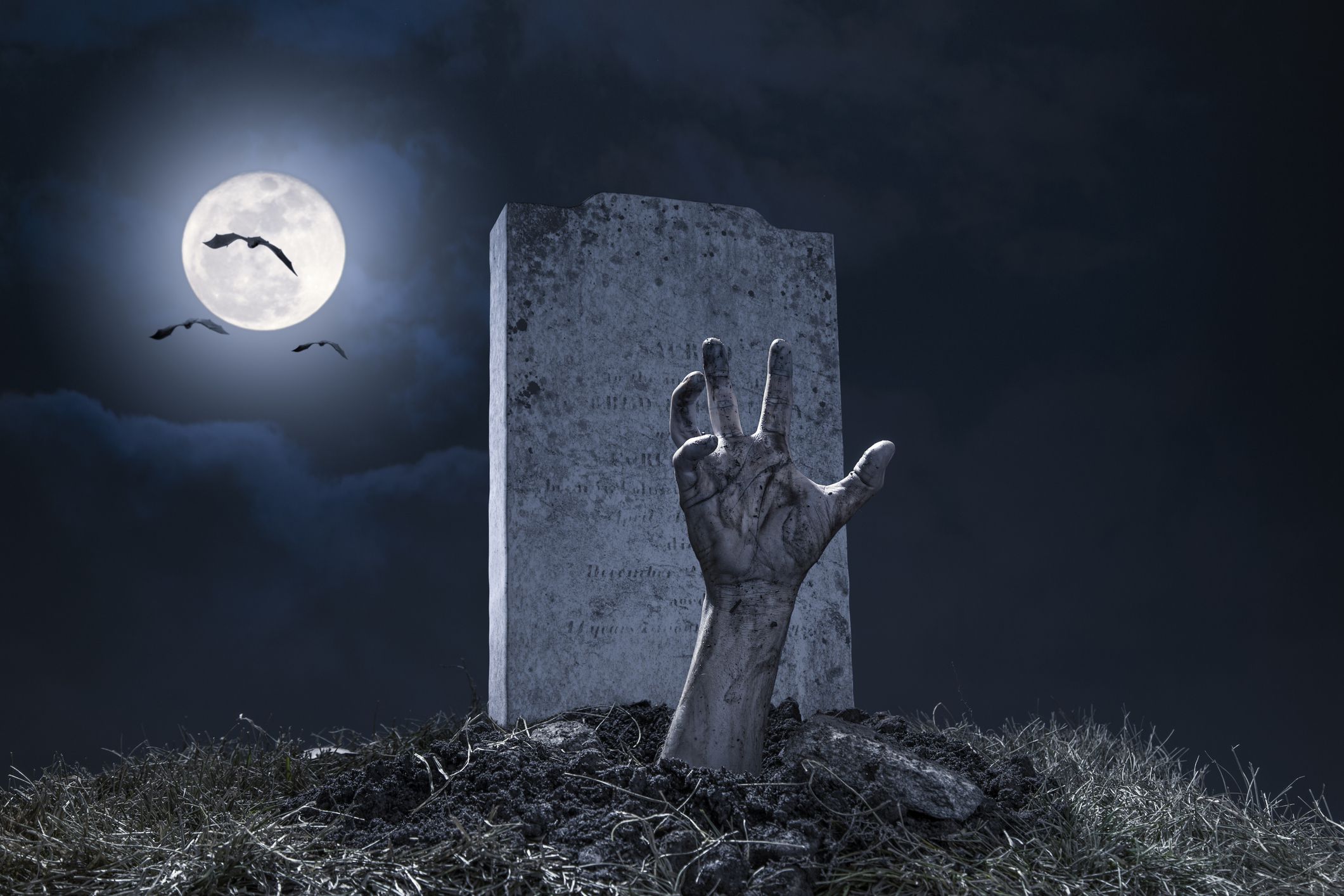 A zombie hand bursting through the grave in a graveyard on Halloween night under a full moon with vampire bats.