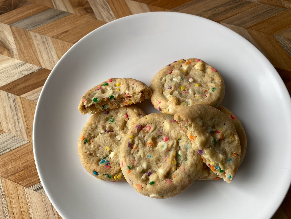 Four funfetti cookies with colorful sprinkles are on a white plate atop a wooden, geometric-patterned table. One cookie is broken in half, showing the soft texture inside.