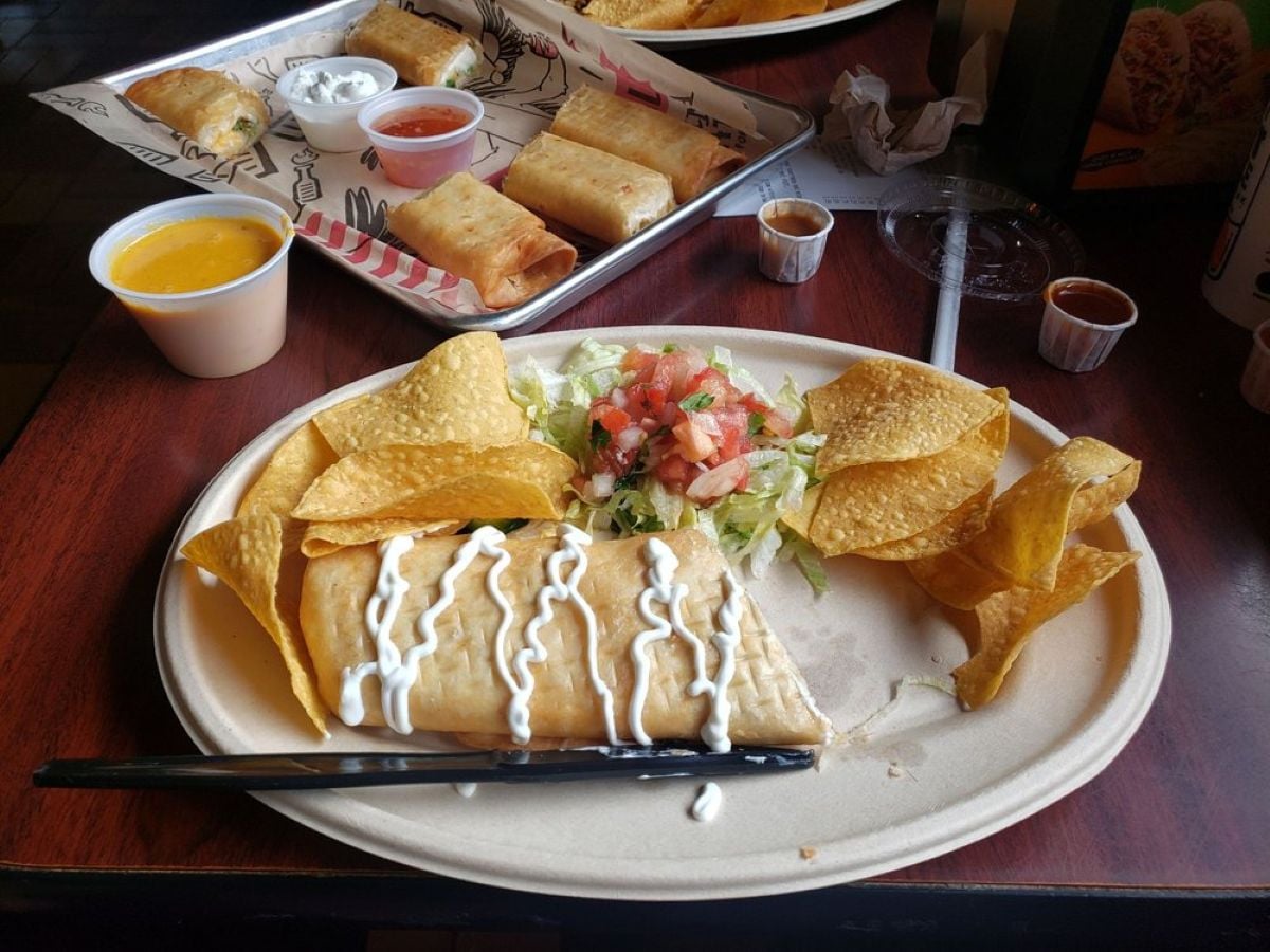 A plate with a chimichanga topped with sour cream, tortilla chips, shredded lettuce, and pico de gallo. In the background are trays with taquitos, dipping sauces, and additional chips on a wooden table.