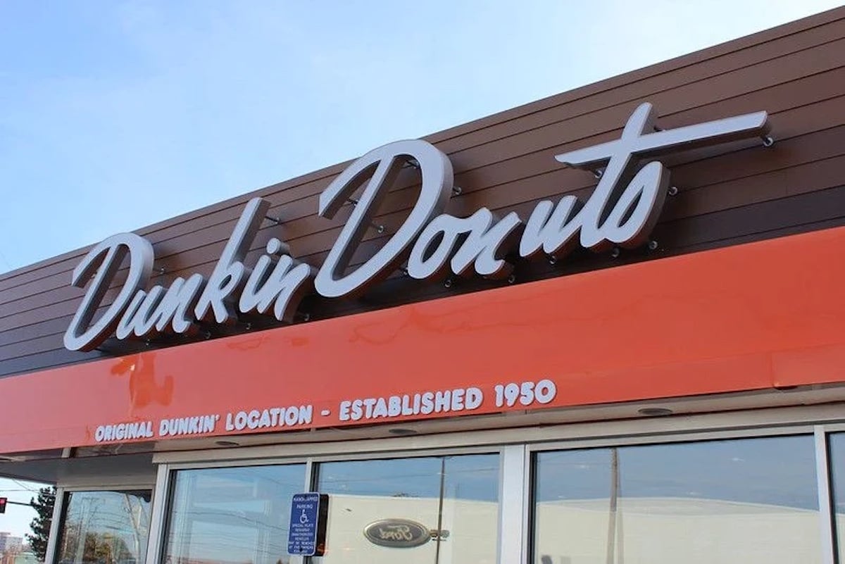 The image shows the exterior of a Dunkin’ Donuts store with a large, cursive sign. Below, an orange awning reads "Original Dunkin' Location – Established 1950," highlighting its place among notable companies founded by year.