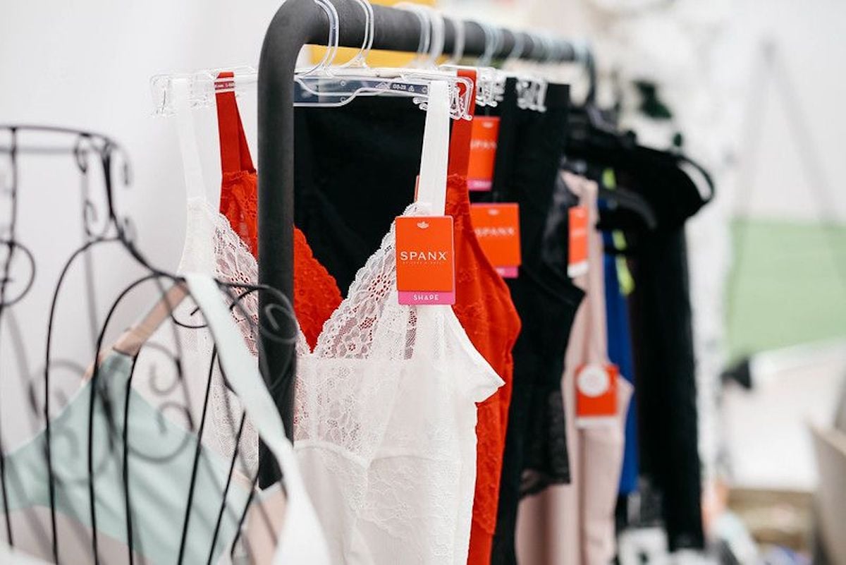 Several SPANX slip dresses in white, red, black, and beige hang on clear hangers with tags attached. Founded in 2000, SPANX is featured here among notable companies founded by year. The background is out of focus.