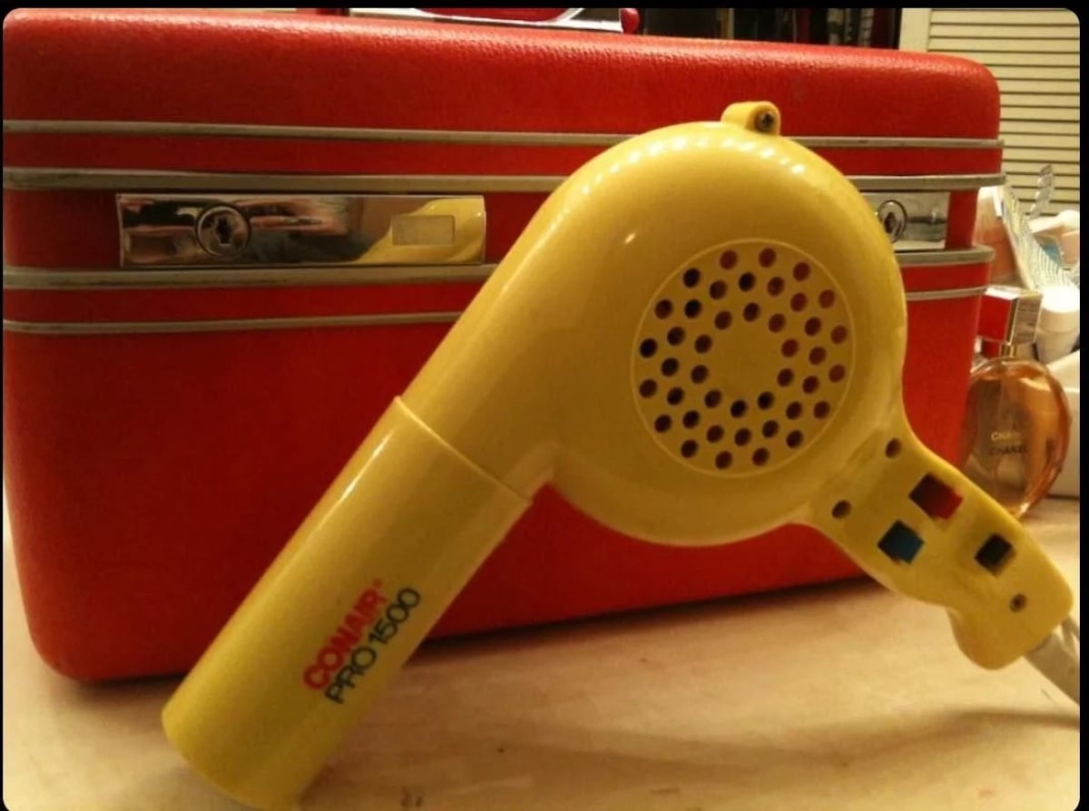 A vintage yellow Conair Pro 1500 hair dryer—by one of the companies founded by year—rests before a bright red suitcase on a countertop, with a small perfume bottle visible to the right.