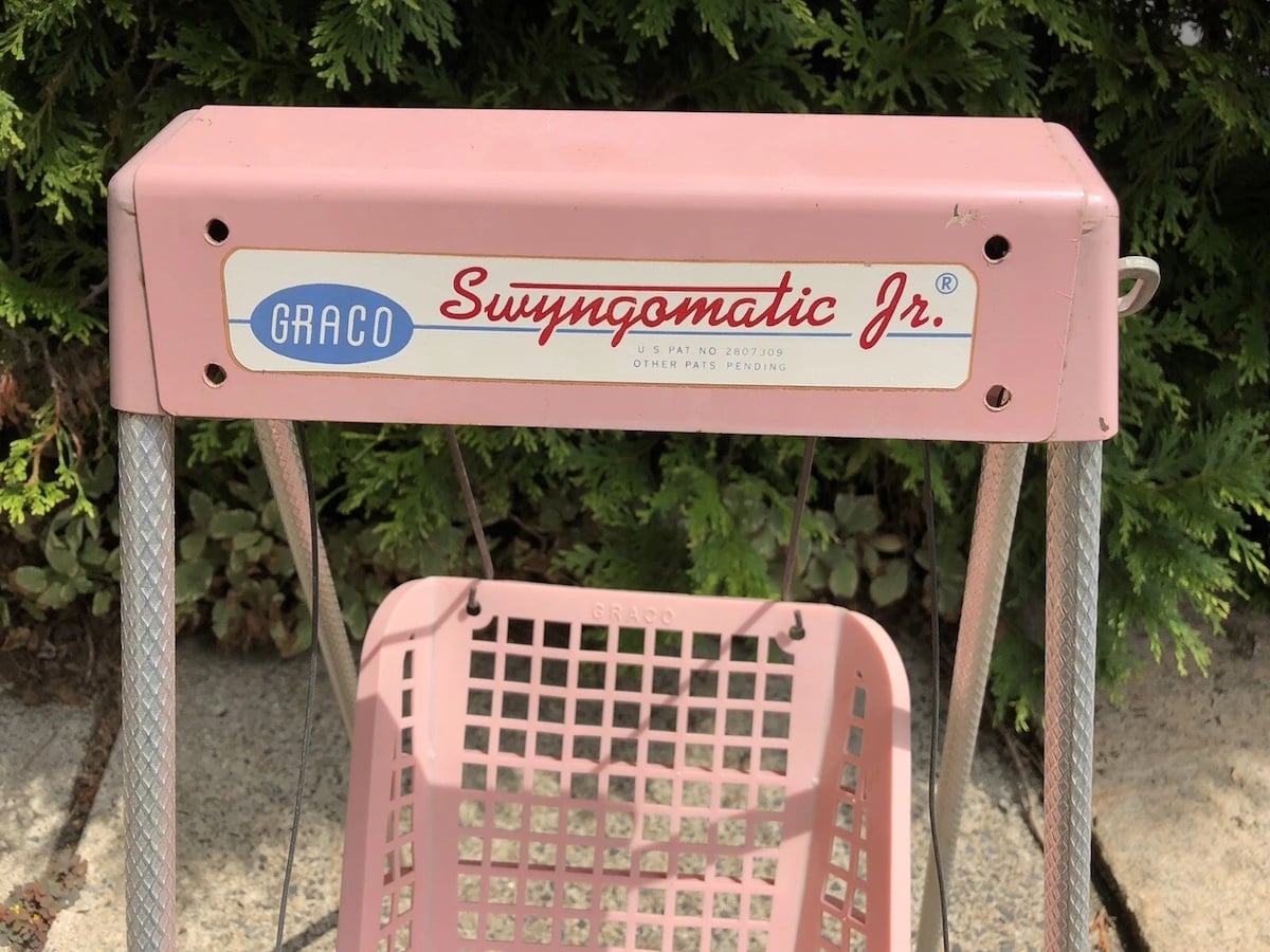 A close-up of a vintage pink Graco Swingomatic Jr. baby swing, from one of the companies founded by year 1942, shows the brand label on top and a perforated plastic seat below, with green bushes in the background.