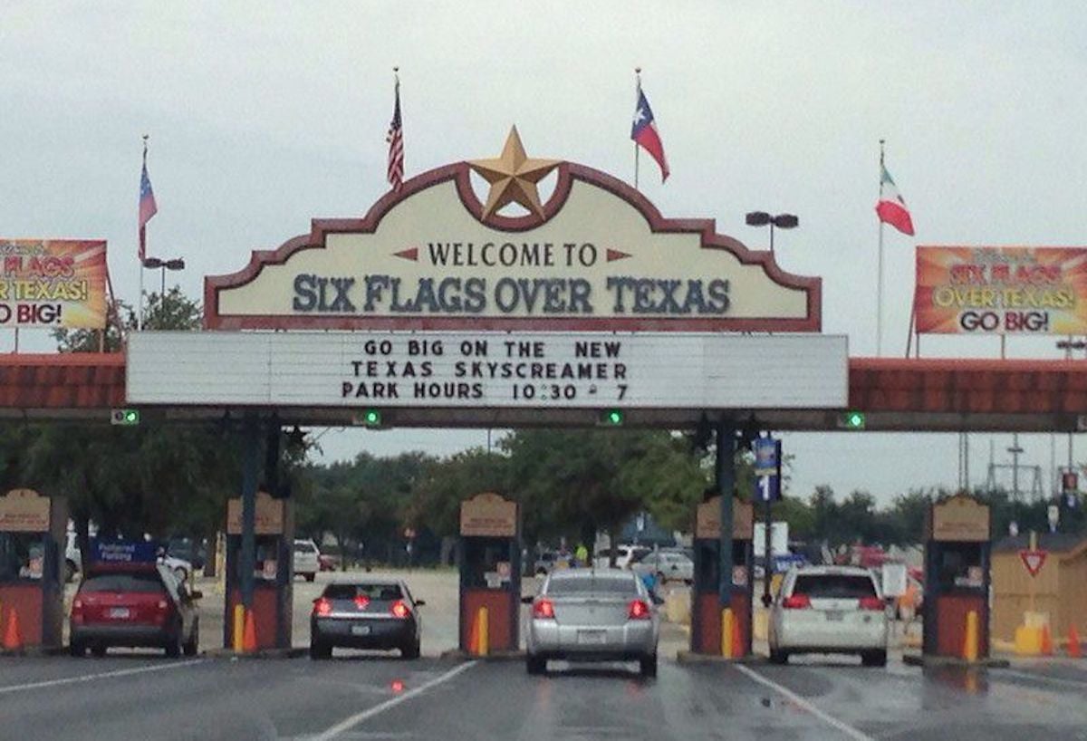 Cars approach the entrance to Six Flags Over Texas, with a large sign displaying park hours and flags above the gate. The sign promotes the new Texas SkyScreamer ride at one of the notable companies founded by year 1961.