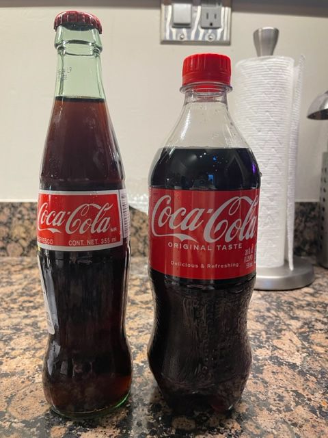 A glass bottle and a plastic bottle of Coca-Cola sit side by side on a kitchen countertop, with a paper towel roll and electrical outlet in the background.