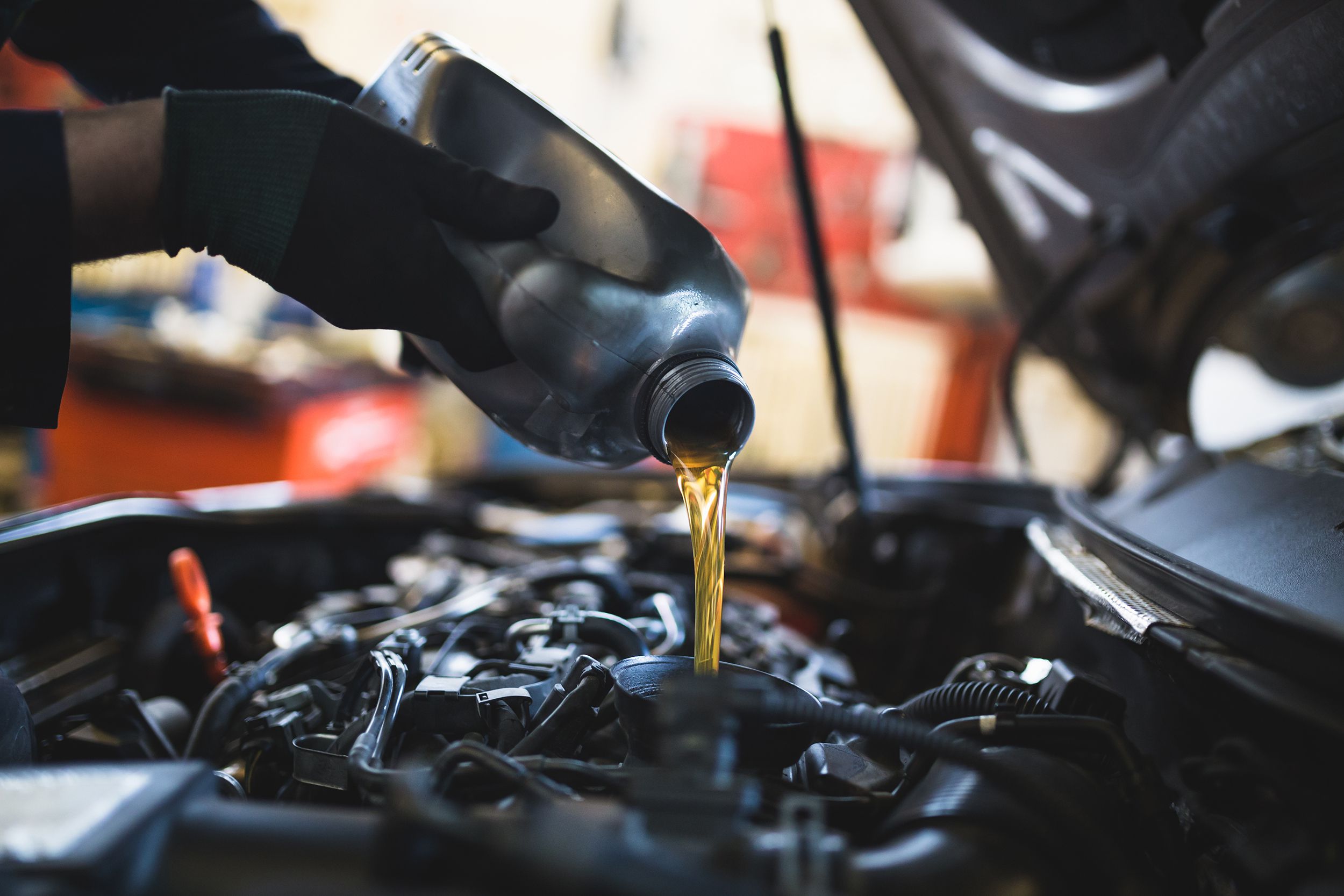 Closeup of car mechanic's hands holding a jug pouring oil under the hood into the engine of a car, selective focus, rest of car engine and repair garage blurred in the background