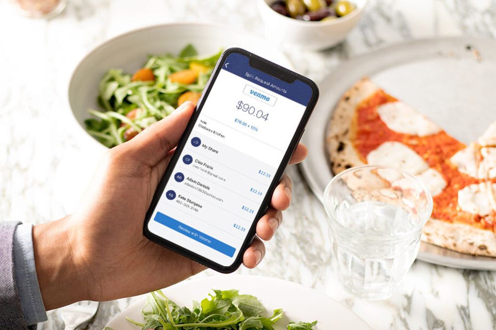 A person holding a smartphone displaying a Venmo balance and payment history at a table with a salad, a plate of pizza, a bowl of olives, and a glass of water.