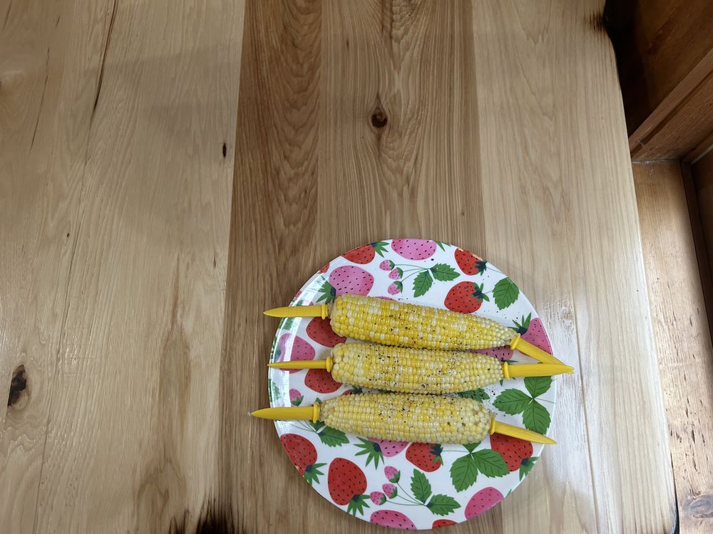 Three ears of corn on the cob with yellow handles are arranged on a colorful plate decorated with strawberries, placed on a light wooden table.