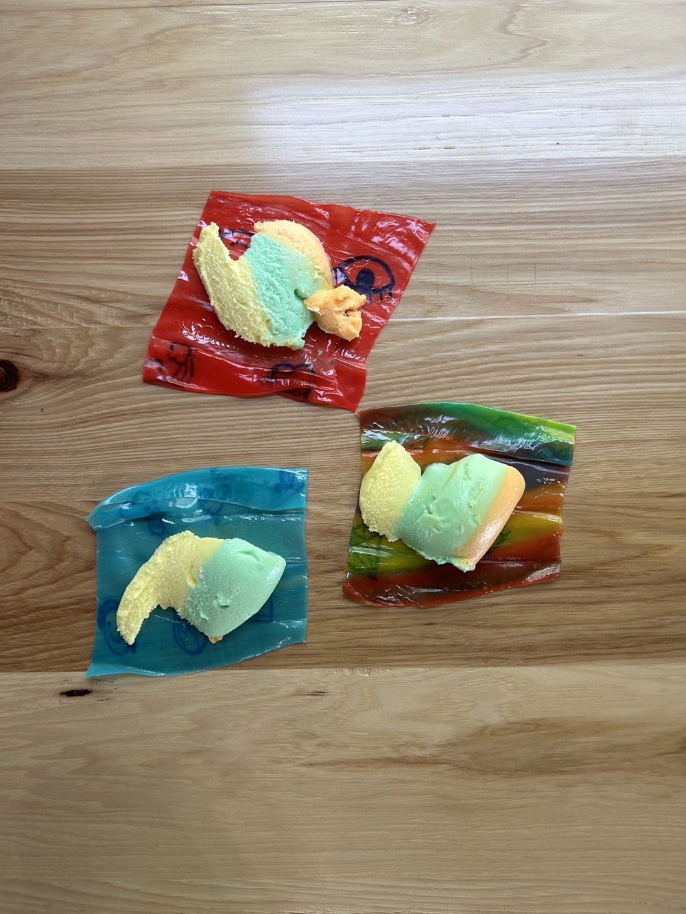 Three pieces of colorful ice cream sit on clear plastic wrappers, arranged on a light wooden surface. The ice cream is pastel yellow, green, and pink, each piece on a different colored wrapper.