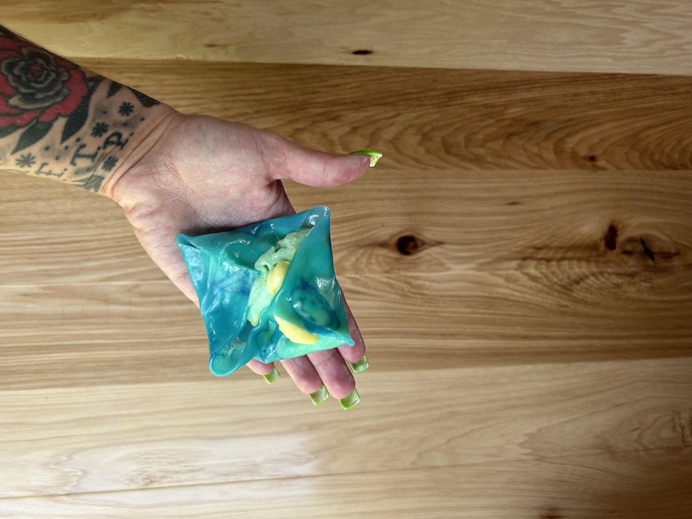 A hand with green nails and a floral tattoo holds a blue, translucent, square-shaped pouch containing yellow and green objects, against a wooden background.