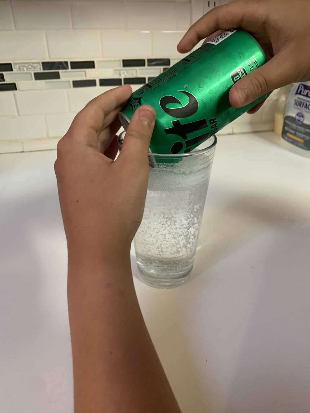 A person pours a can of 7UP soda into a clear glass on a white kitchen counter with white tile backsplash.