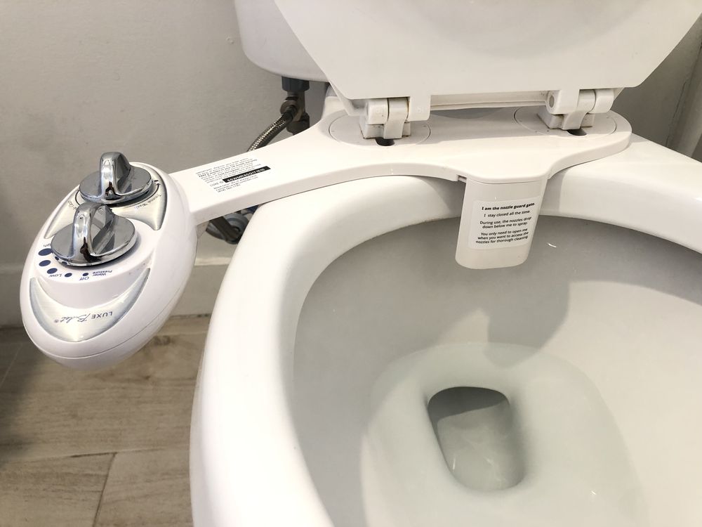 A close-up of a white toilet with an attached bidet system featuring two control knobs on the left side. The toilet is clean, and the bidet attachment is positioned under the seat.