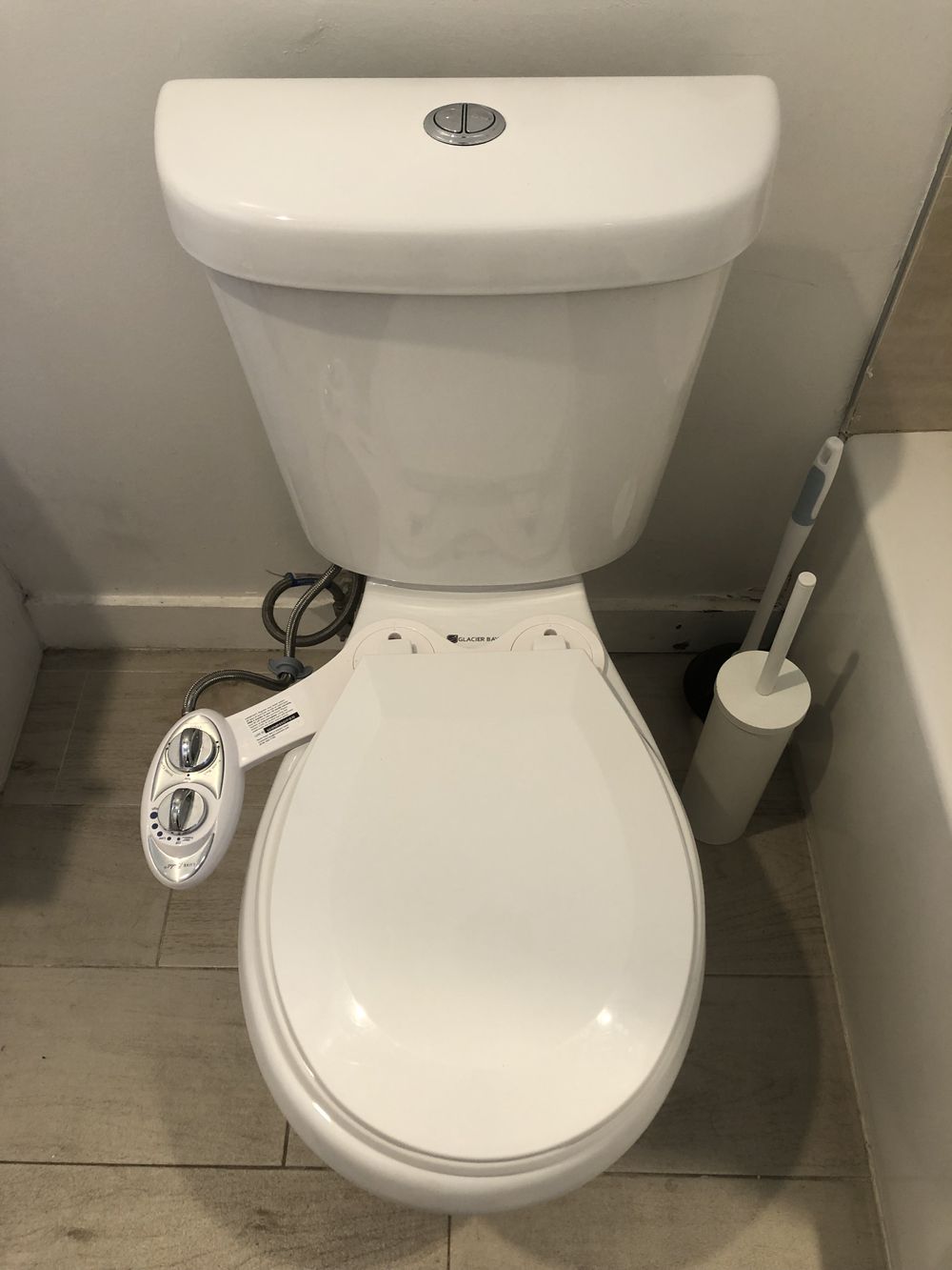 A white toilet with a bidet attachment featuring control knobs on the left side, next to a white toilet brush and holder, set on light-colored tiled flooring.