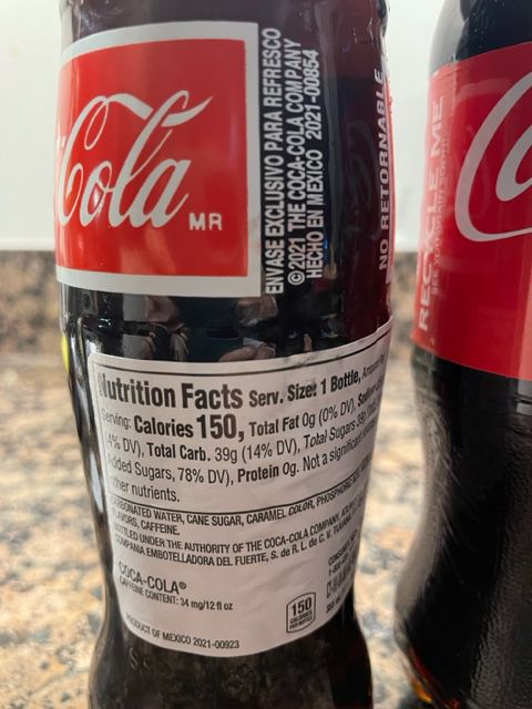 A close-up of a Coca-Cola glass bottle label showing nutrition facts, including 150 calories per serving and 39g of total sugars. The label also contains ingredients and recycling information in English and Spanish.