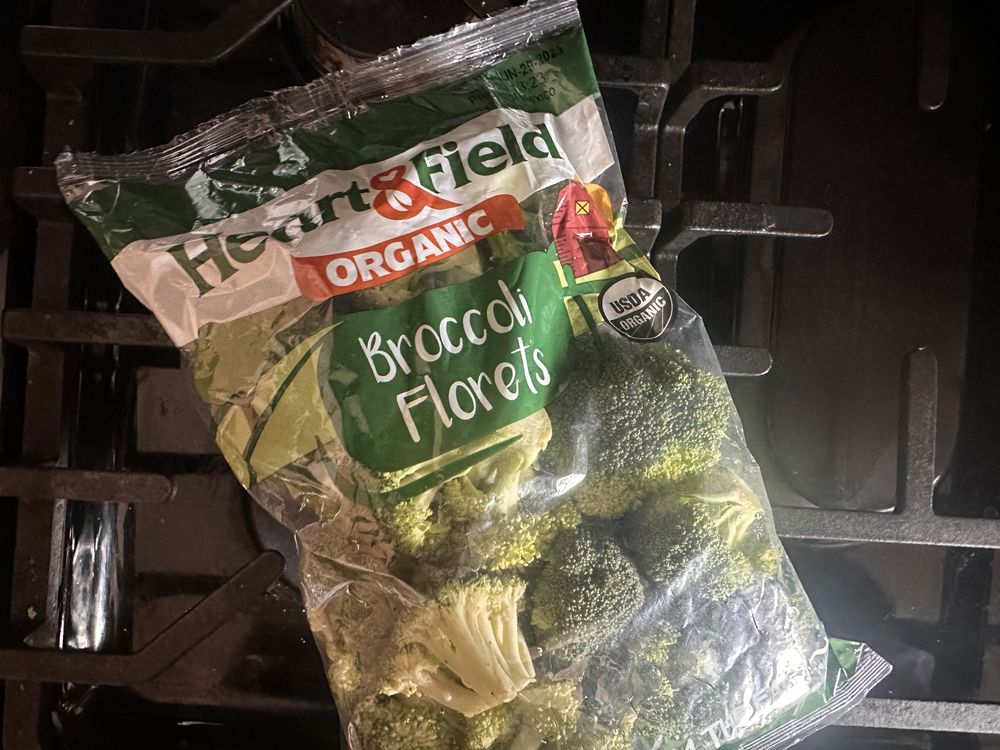 A bag of Heart & Field Organic Broccoli Florets sits on a black stovetop. The bag is partially open, revealing fresh green broccoli inside. The packaging displays 