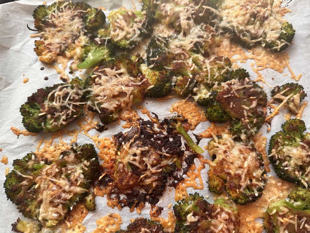 Roasted broccoli slices on parchment paper, topped with melted and slightly browned shredded cheese. Some pieces appear crispy and caramelized around the edges.