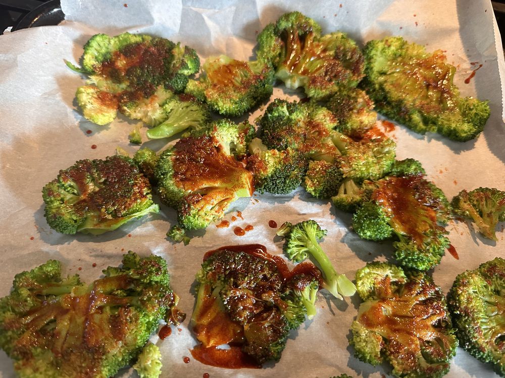 Broccoli florets spread on parchment paper, topped with a reddish-brown sauce and roasted, showing slightly charred and crispy edges.