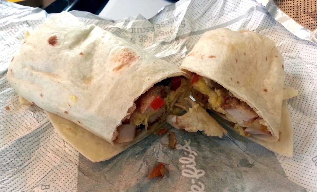 A breakfast burrito cut in half reveals scrambled eggs, potatoes, sausage, and red peppers inside—capturing the classic appeal of the best fast-food breakfast burrito, served on crumpled paper wrapping.