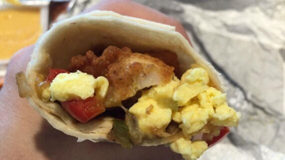 A close-up of a hand holding one of the best fast-food breakfast burritos, packed with scrambled eggs, fried chicken, peppers, and melted cheese, with a foil wrapper visible in the background.
