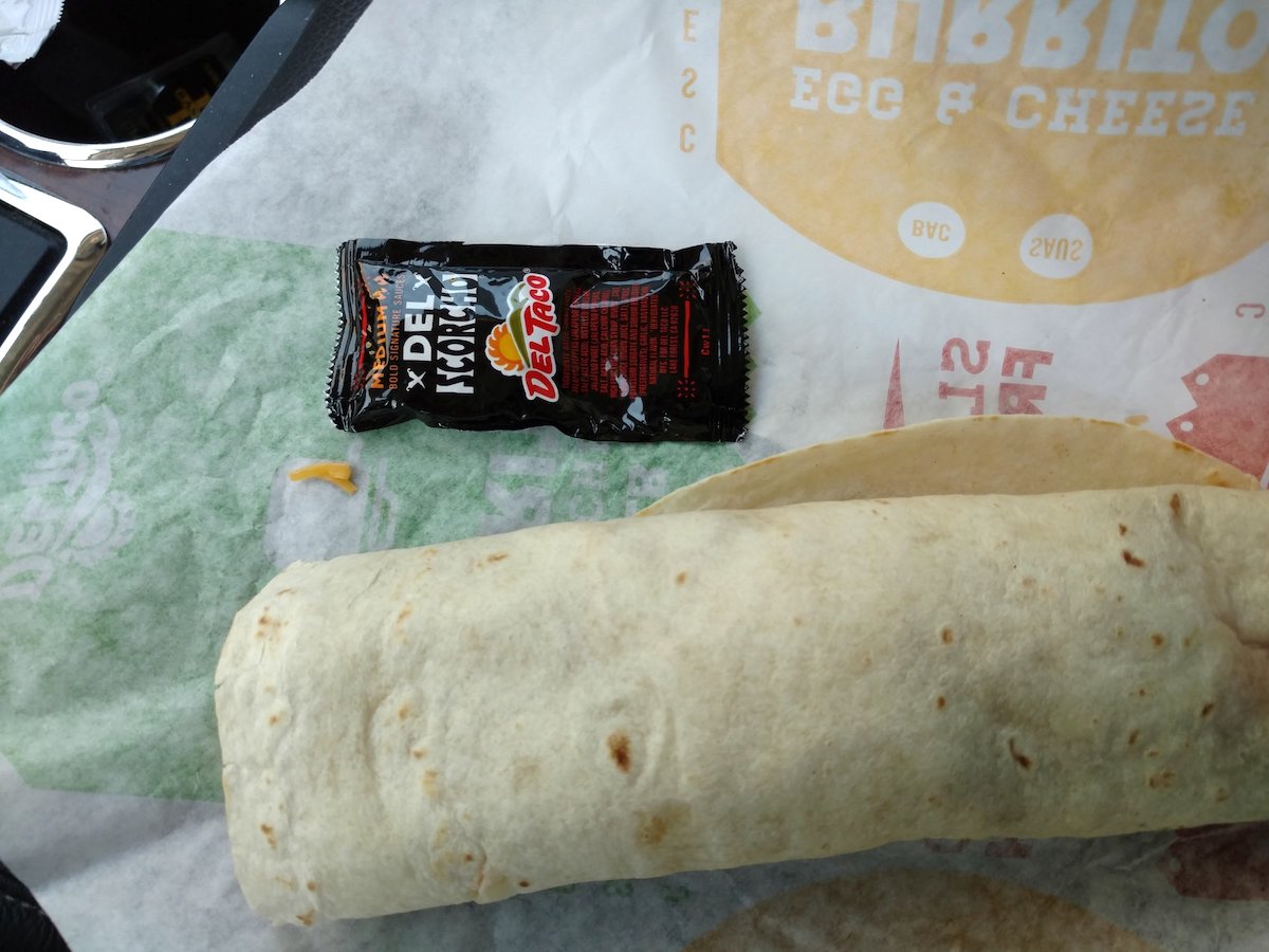 A wrapped burrito sits on a Taco Bell wrapper next to a packet of Taco Bell Diablo hot sauce, on a car seat—ready to be enjoyed as one of the best fast-food breakfast burritos around.