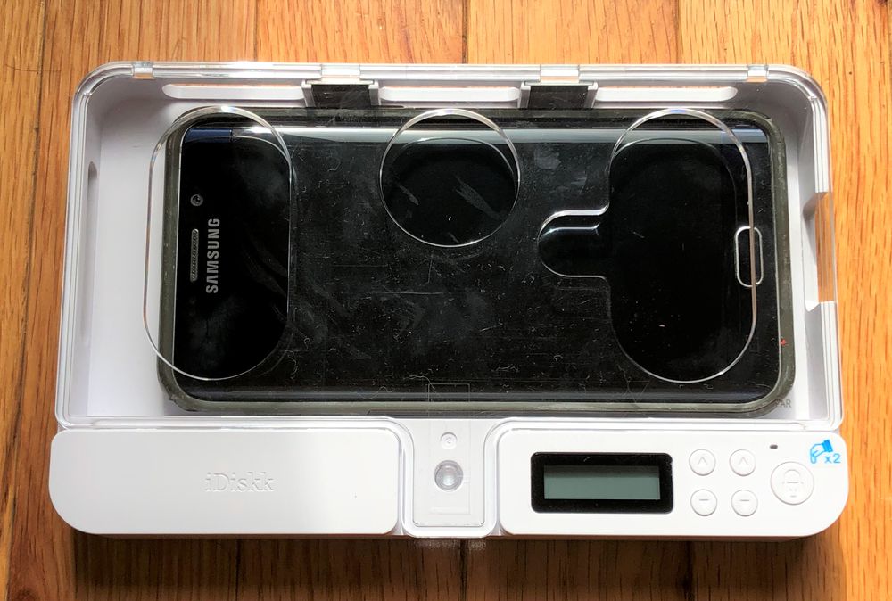 A Samsung smartphone inside a white electronic device with a transparent lid, circular cutouts, a small display screen, and control buttons, placed on a wooden surface.